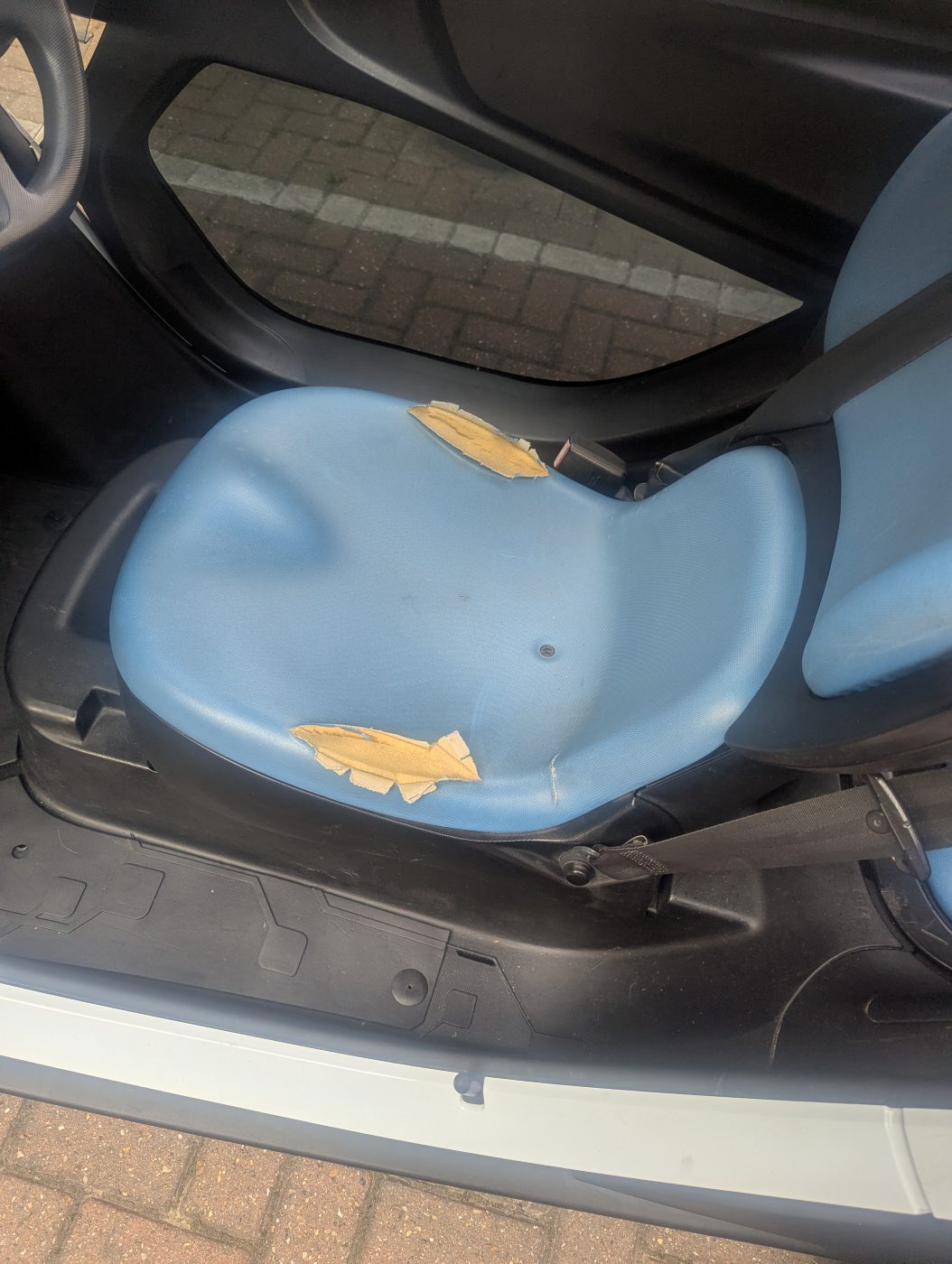 The driver's seat base, showing two torn areas in the blue fabric with the foam
                underneath exposed